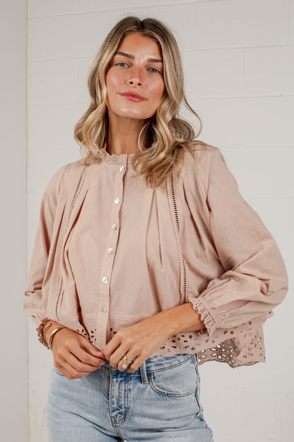 A woman wearing a dusty rose button-up blouse with eyelet lace details and light blue jeans, standing against a white brick wall.