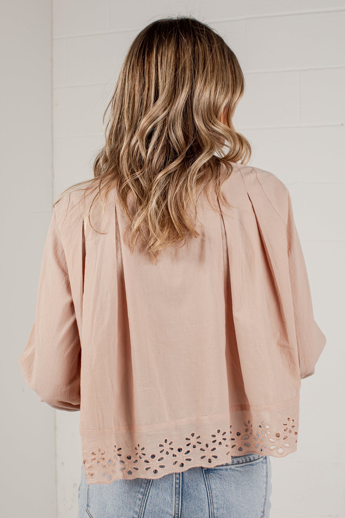 A woman viewed from behind wearing a dusty pink blouse with pleated shoulders and eyelet lace trim at the hem, paired with light blue jeans against a white brick wall.