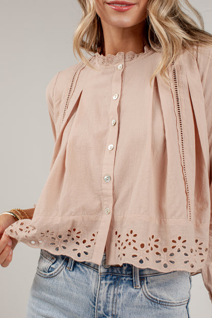 A woman wearing a light blush pink blouse with eyelet lace trim at the collar and hem, button-front closure, and pleated side details, paired with light-wash blue jeans against a plain light gray studio background.