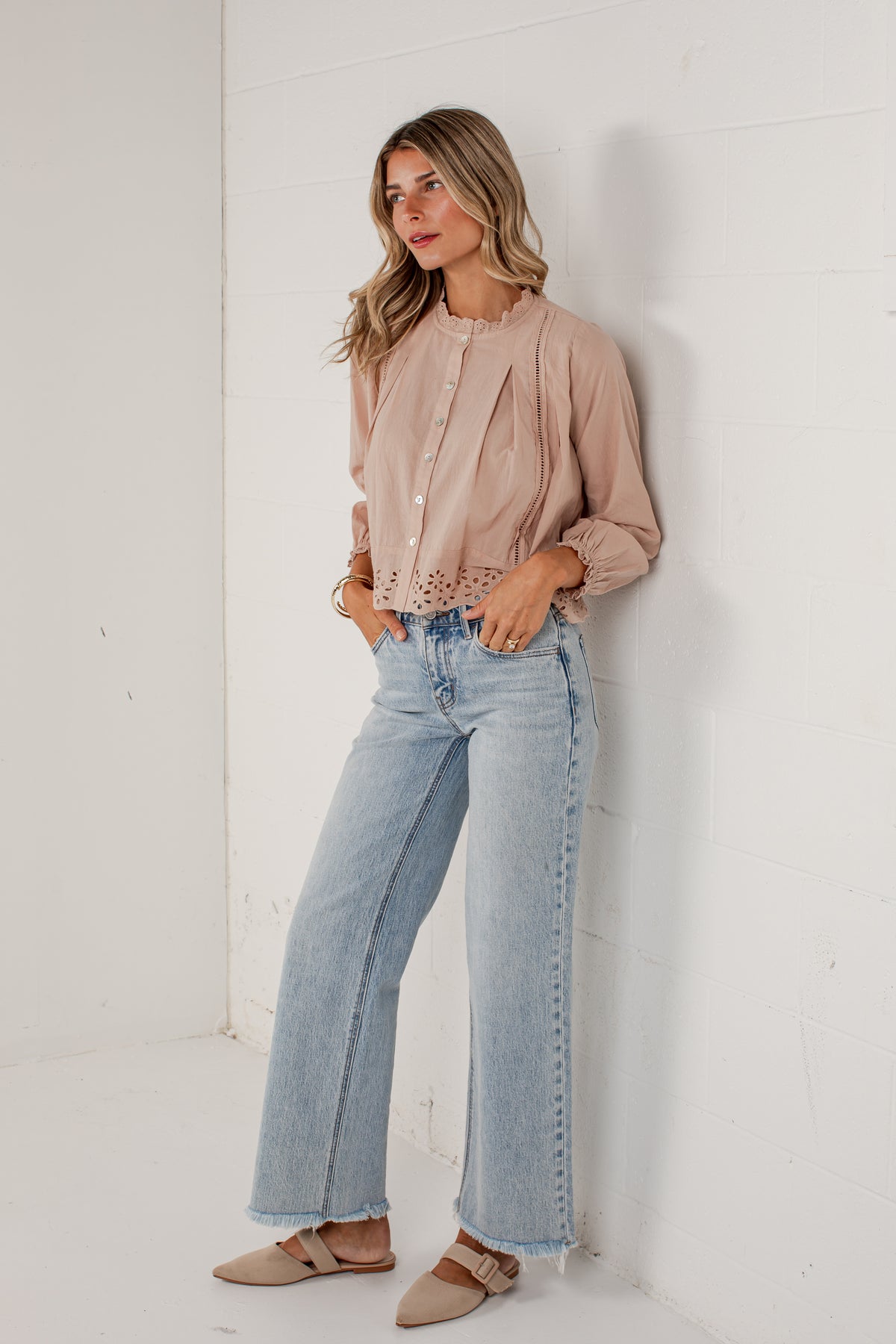 A woman stands against a white wall wearing a light blush pink long-sleeve blouse with lace-up front and eyelet cutouts, light-wash wide-leg blue jeans, and beige flat sandals.