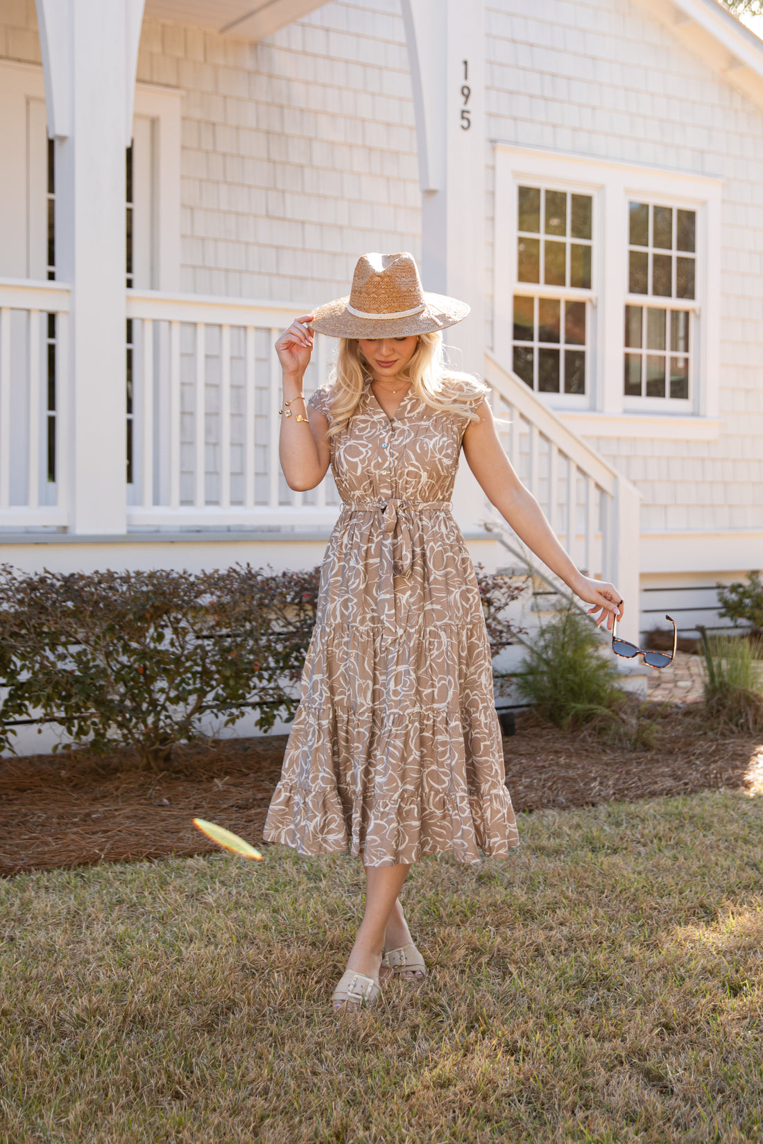 Woman in a floral dress and hat standing in front of a white house.
