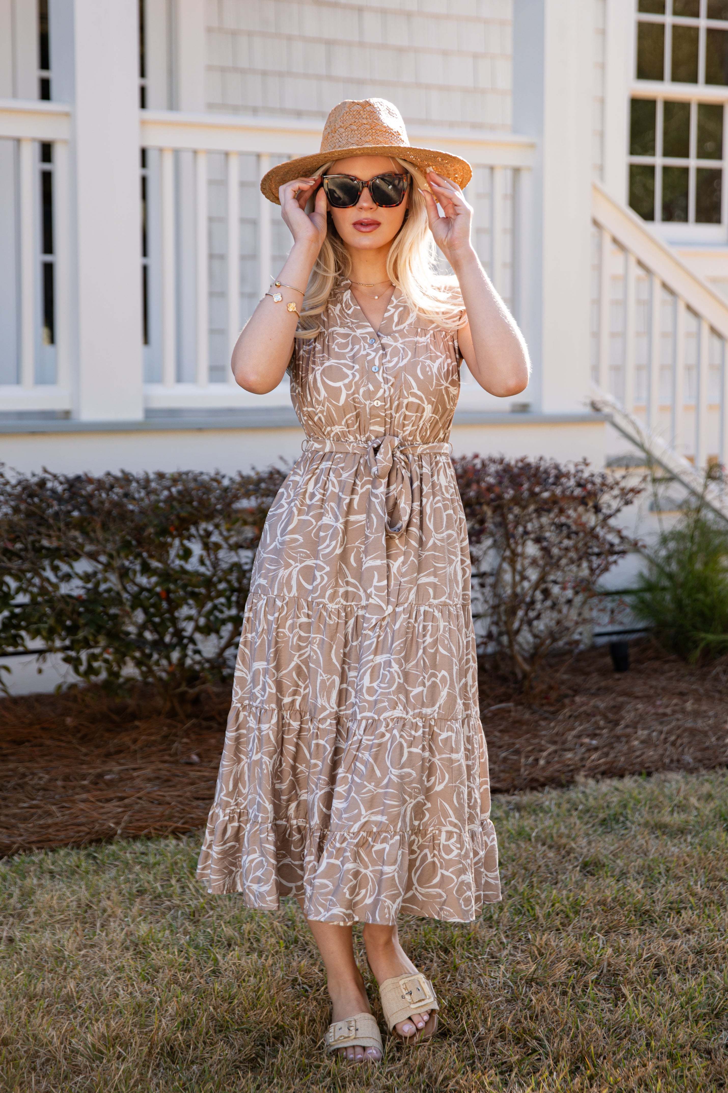 Woman in a patterned dress and straw hat standing in front of a white house.
