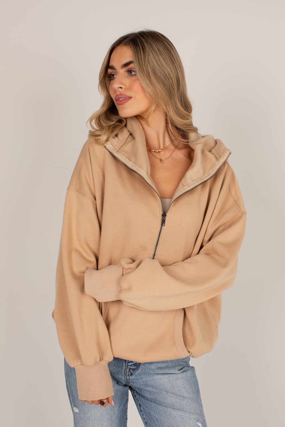 A woman wearing a camel-colored half-zip hoodie with a hood and voluminous sleeves, paired with light-wash blue jeans, photographed against a neutral light gray studio background.