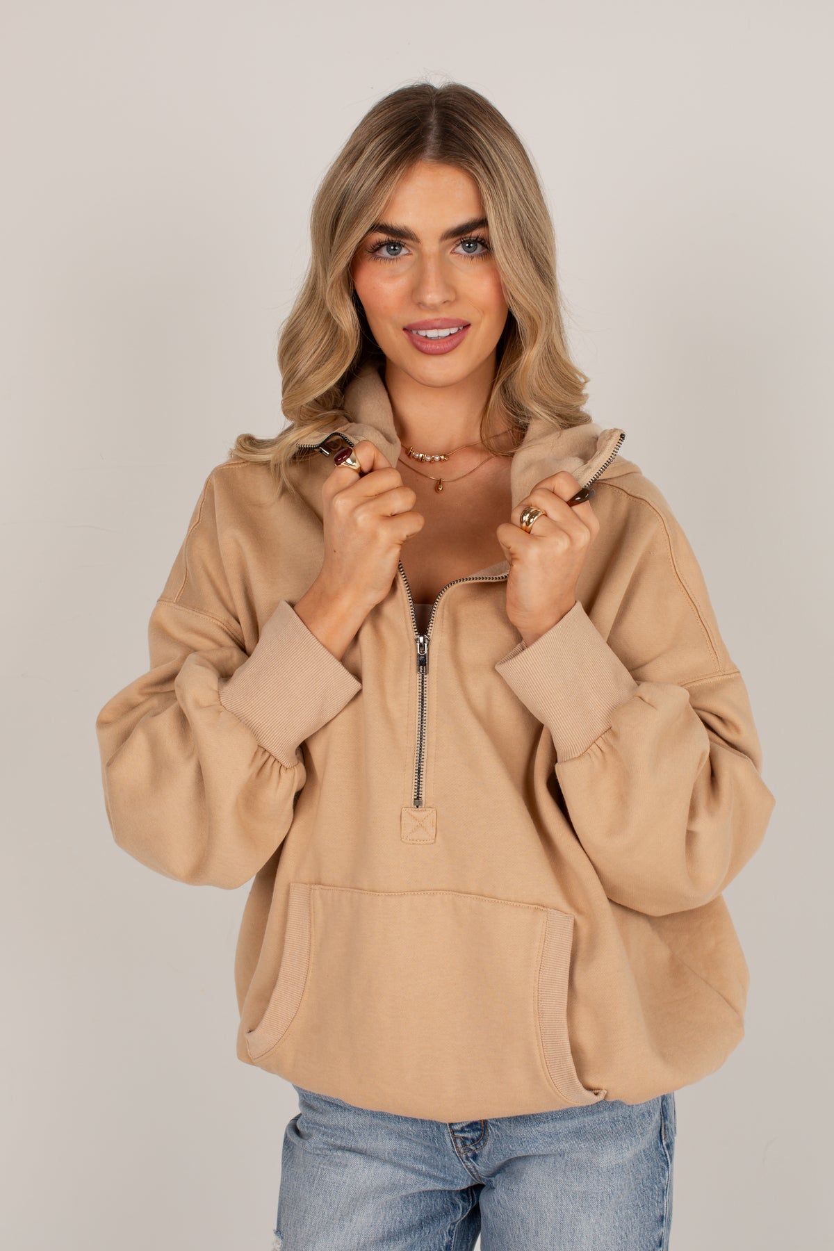 A woman wearing a camel-colored half-zip hoodie with a kangaroo pocket and voluminous sleeves, paired with blue jeans, against a plain light gray background.