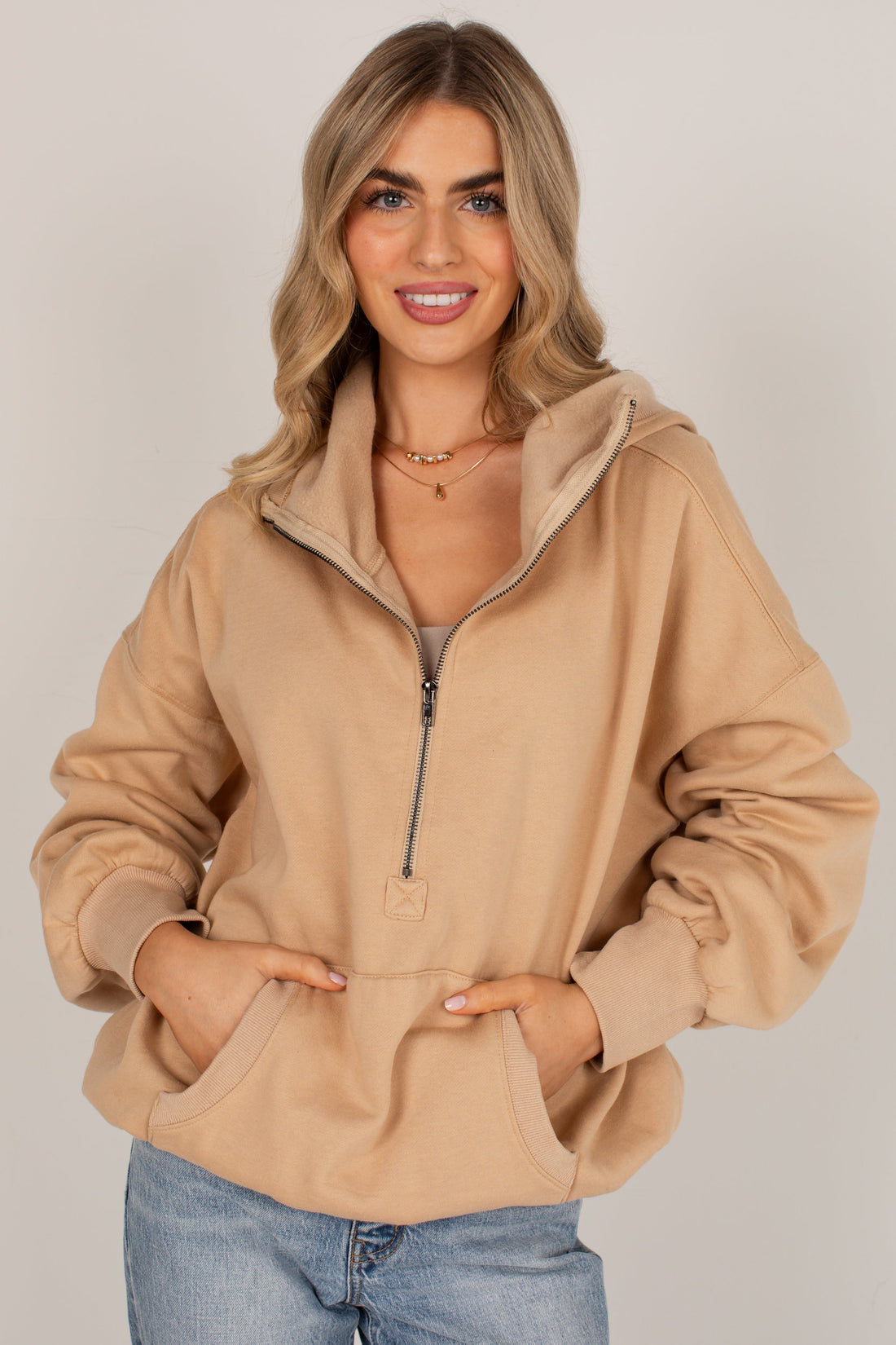A woman modeling a beige half-zip hoodie with a drawstring hood and front pockets, paired with light blue jeans, against a plain light gray studio background.