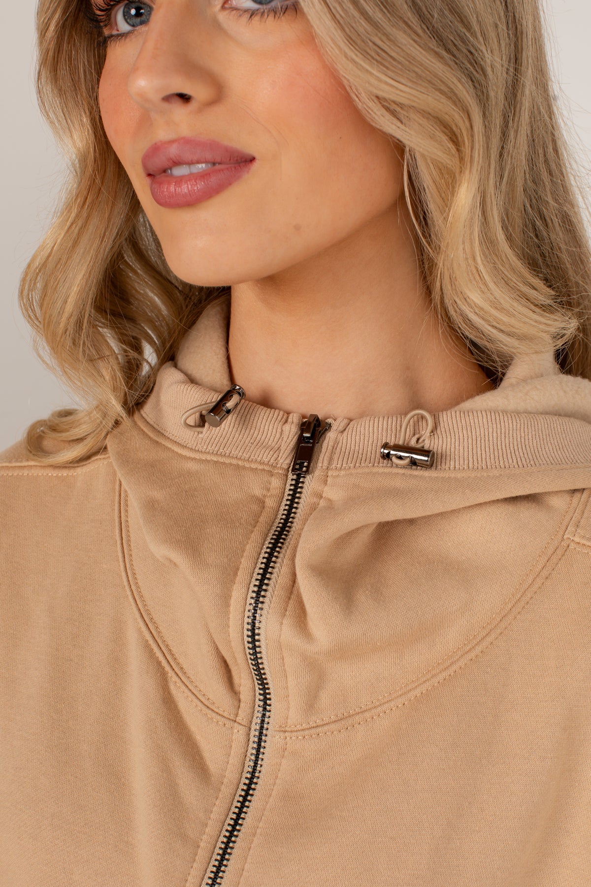Close-up of a woman wearing a beige zip-up hoodie with a ribbed hood and drawstrings, shown against a light neutral background.