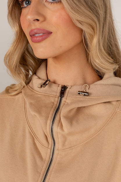 Close-up of a woman wearing a beige zip-up hoodie with a ribbed hood and drawstrings, shown against a light neutral background.