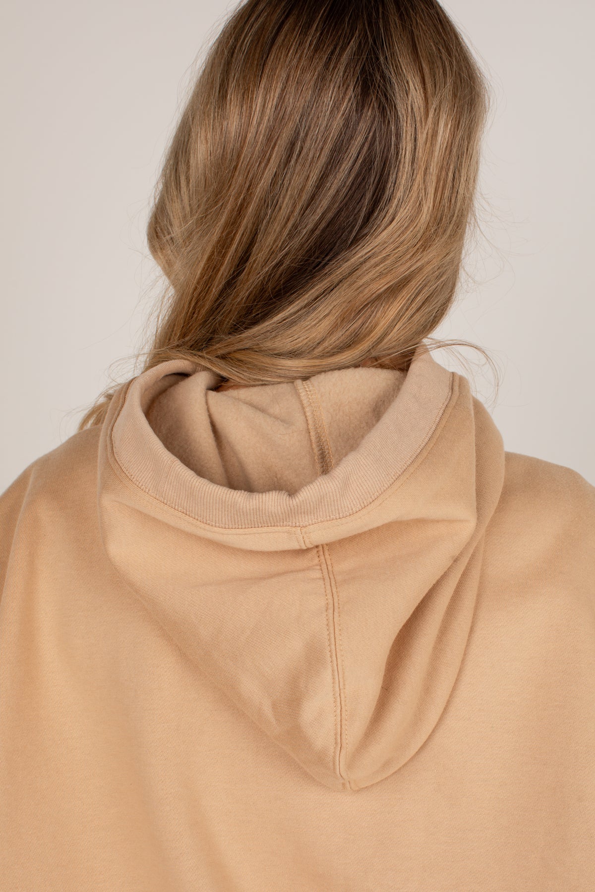 Back view of a person wearing a light tan or beige pullover hoodie, showing the hood and upper back against a plain light background.