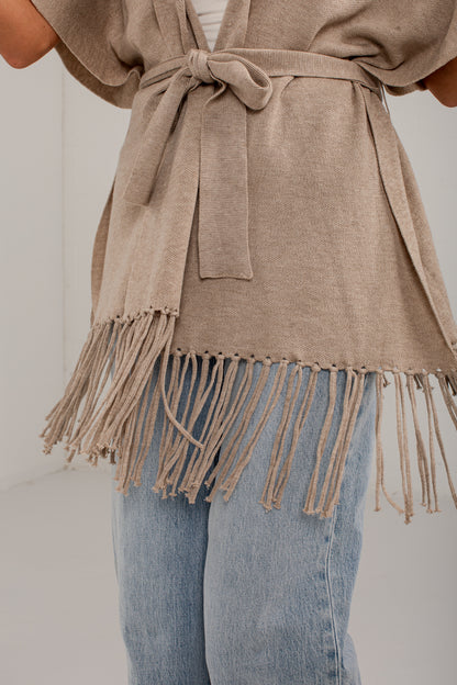 A close-up of a person wearing a taupe knit wrap cardigan with a tie waist and fringe hem, paired with light blue jeans.