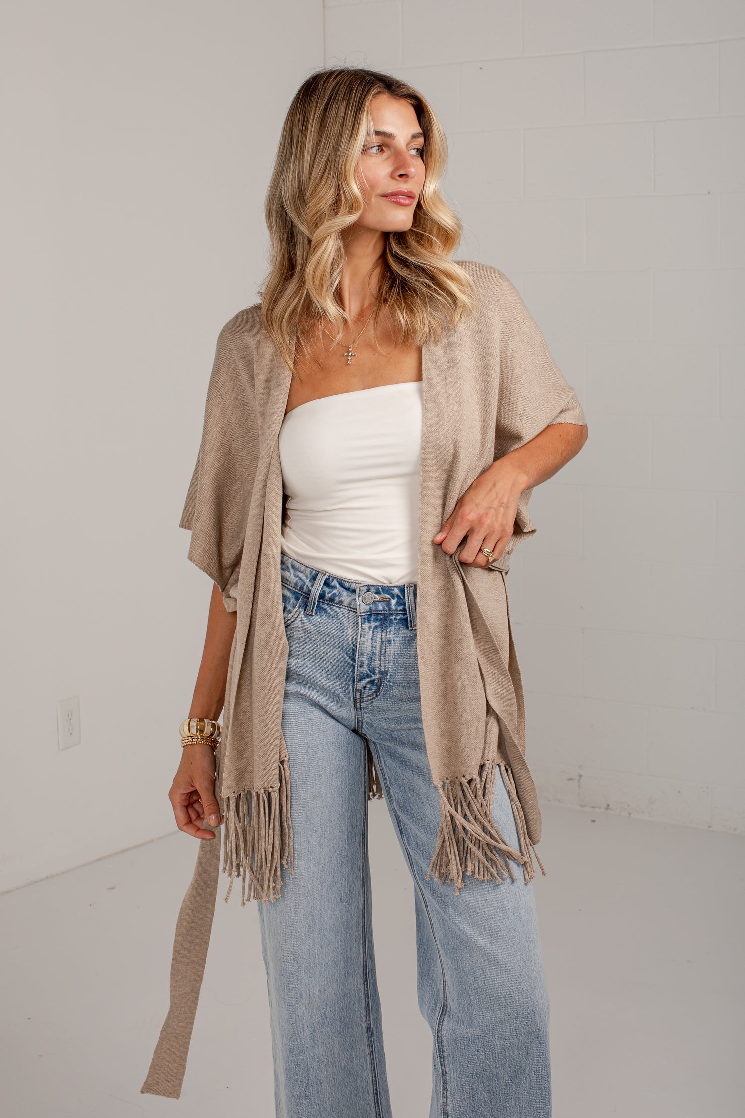 A woman models a light beige open-front fringed cardigan over a white tube top and light-wash blue jeans, standing against a white wall.