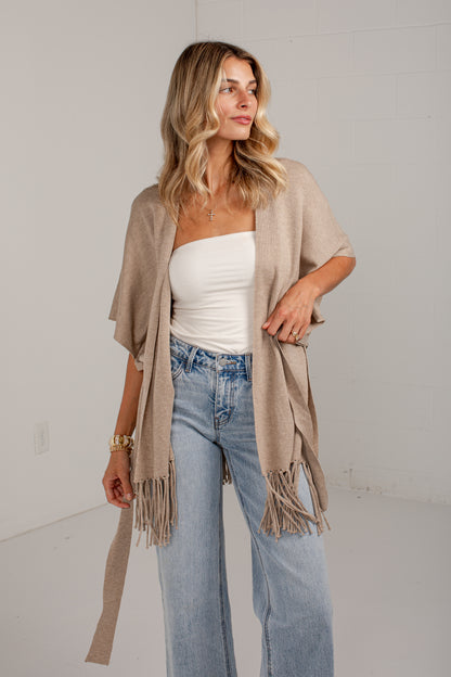A woman models a light beige open-front fringed cardigan over a white tube top and light-wash blue jeans, standing against a white wall.
