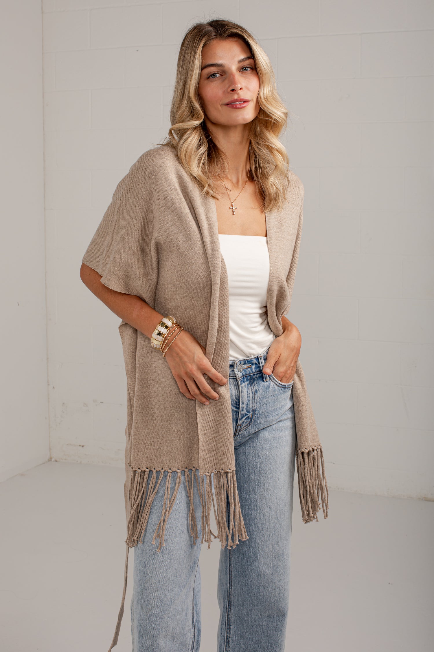 A woman modeling a light beige open-front fringed cardigan over a white tube top and light blue jeans, standing against a white wall.