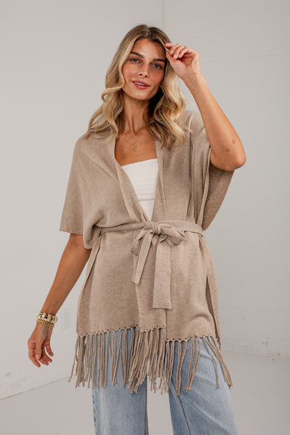 A woman models a light taupe knit wrap cardigan with fringe trim and a tie waist belt, worn over a white top and light blue jeans against a plain white studio background.