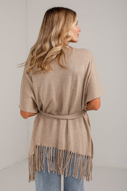 A woman shown from the back wearing a light taupe-beige belted poncho top with fringe trim, paired with blue jeans, in a bright studio setting.