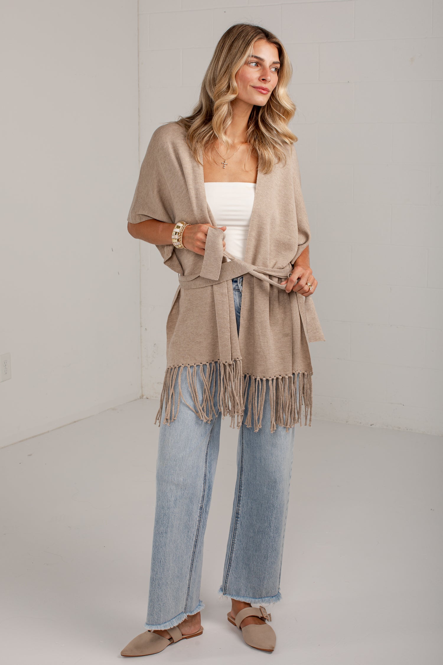 A woman models a light beige knit wrap cardigan with fringe trim, tied at the waist over a white tube top and light blue wide-leg jeans.