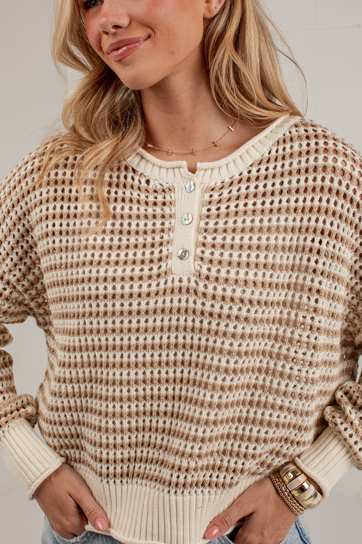 A woman wearing a beige and brown waffle-knit henley sweater with a buttoned collar, paired with light blue jeans.
