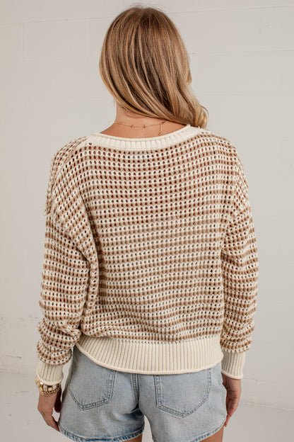 A woman viewed from behind wearing a cream and brown waffle-knit sweater with light blue denim shorts, standing against a white wall.