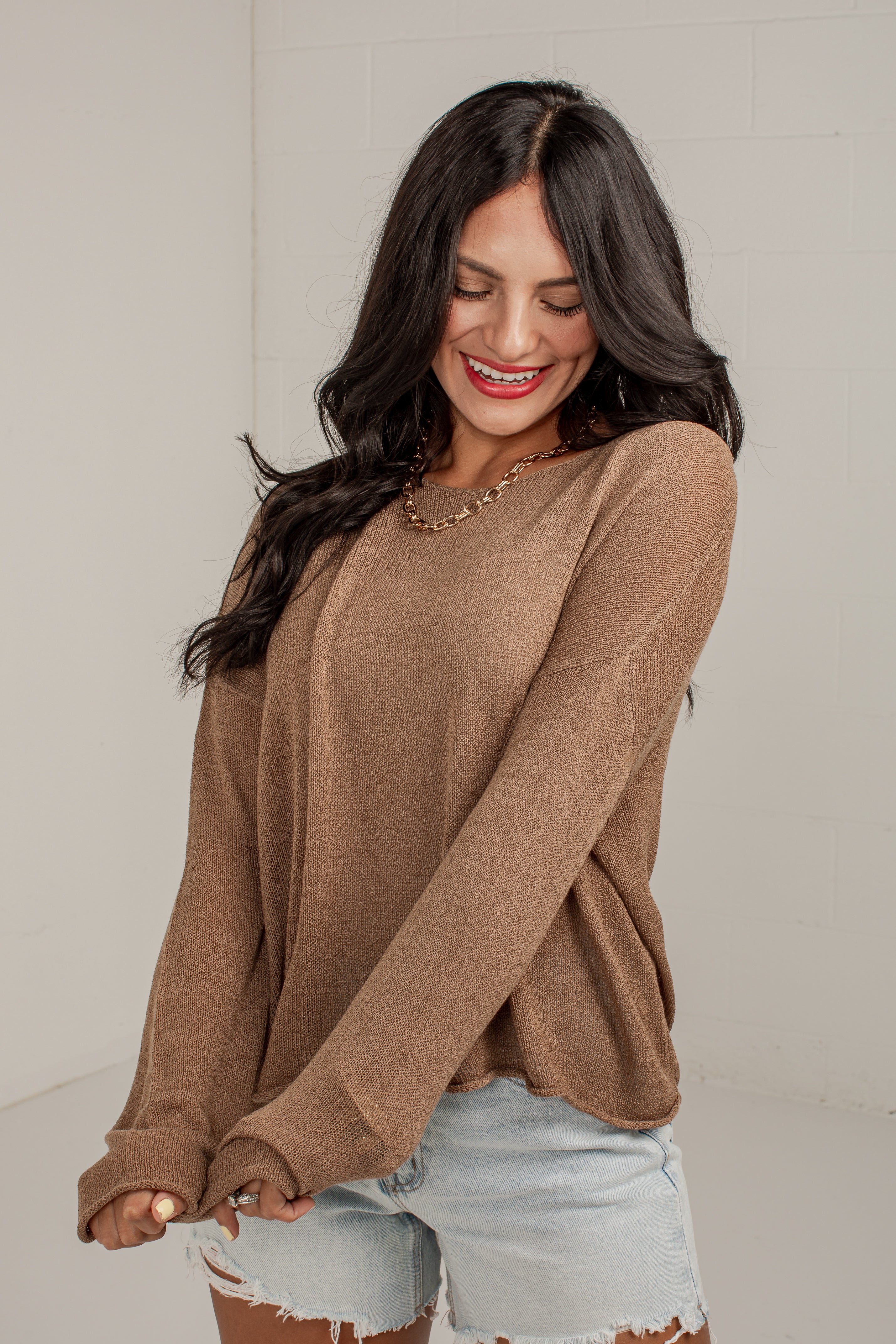 A woman wearing a brown long-sleeve knit sweater and light-wash denim shorts, standing against a light gray wall.