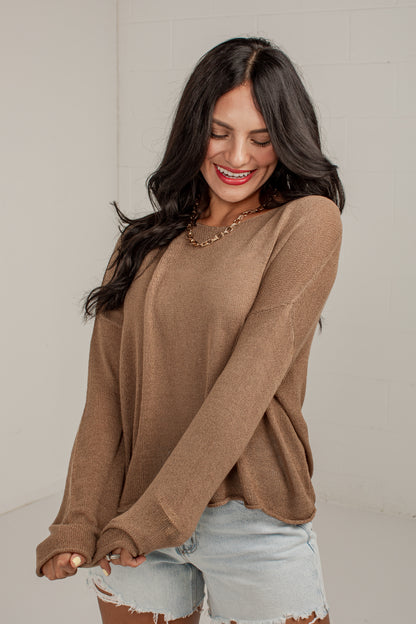 A woman wearing a brown long-sleeve knit sweater and light-wash denim shorts, standing against a light gray wall.