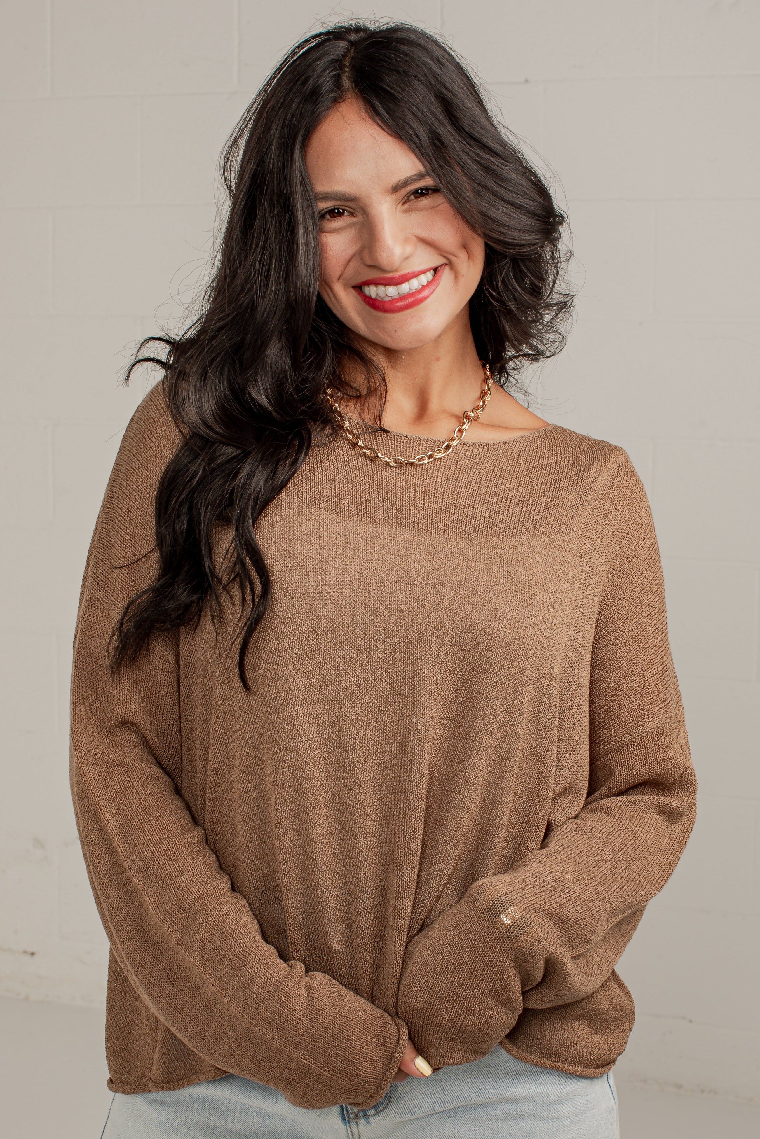A woman with long dark hair smiling while wearing a loose-fitting, long-sleeve knit sweater in a warm taupe-brown color, paired with light-wash denim shorts.