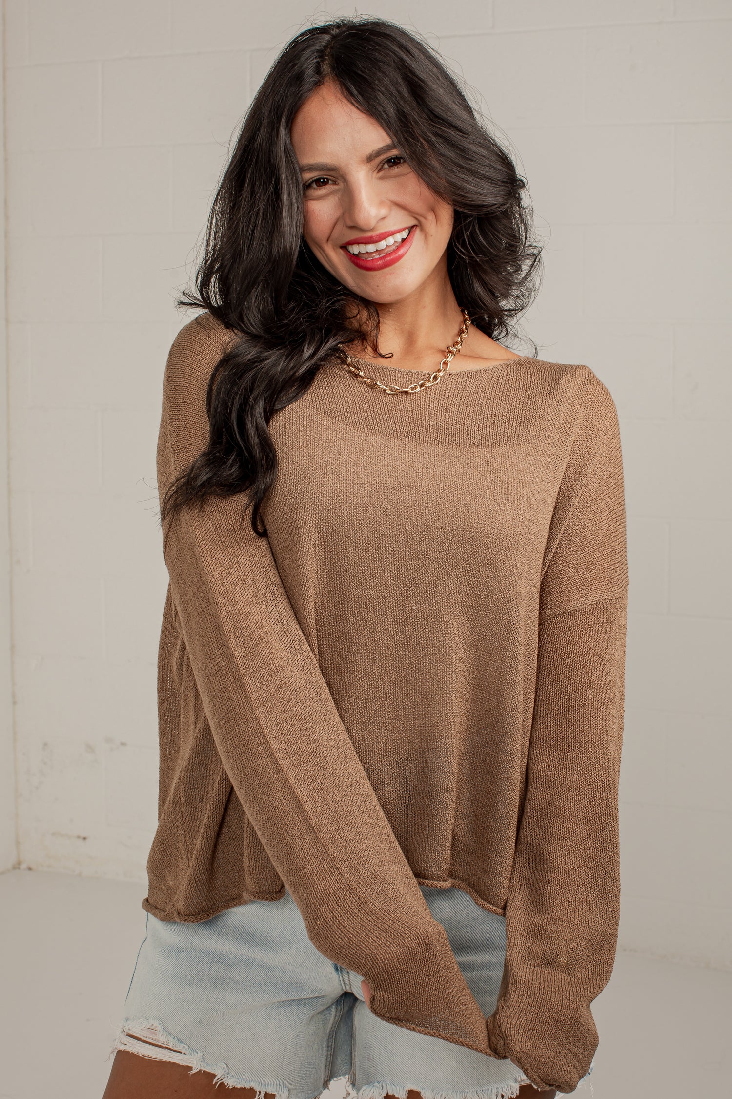 A woman wearing a loose, long-sleeve knit sweater in a warm taupe-brown color, paired with light-wash denim shorts, standing against a light-colored brick wall.