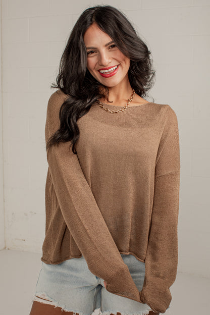 A woman wearing a loose, long-sleeve knit sweater in a warm taupe-brown color, paired with light-wash denim shorts, standing against a light-colored brick wall.