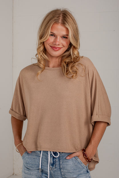 A woman with blonde hair wearing a beige short-sleeve cropped t-shirt and blue jeans, standing against a white brick wall.