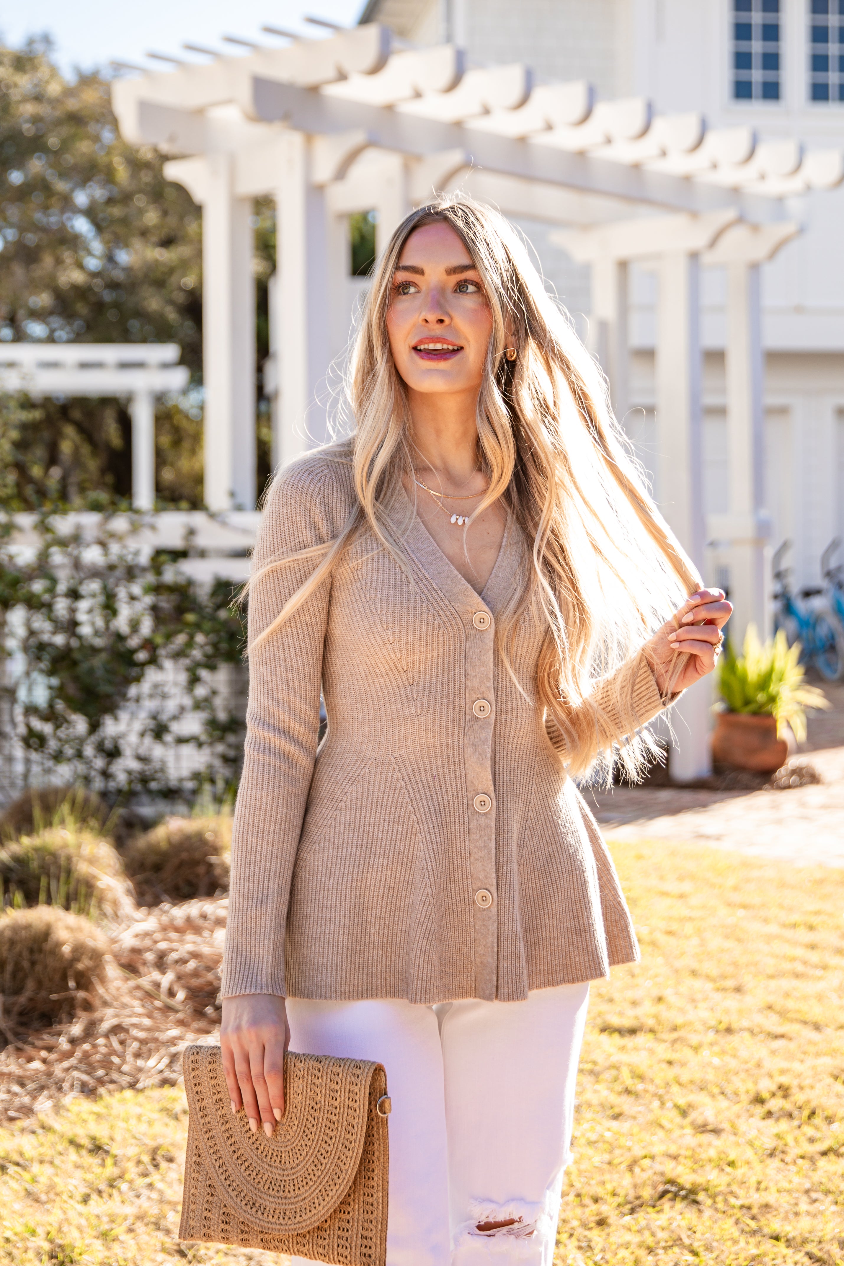 Woman wearing a beige cardigan outdoors with a white building and greenery in the background
