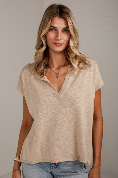 A woman wearing a beige short-sleeve V-neck top with a textured knit fabric, paired with light blue jeans, standing against a light gray brick wall.