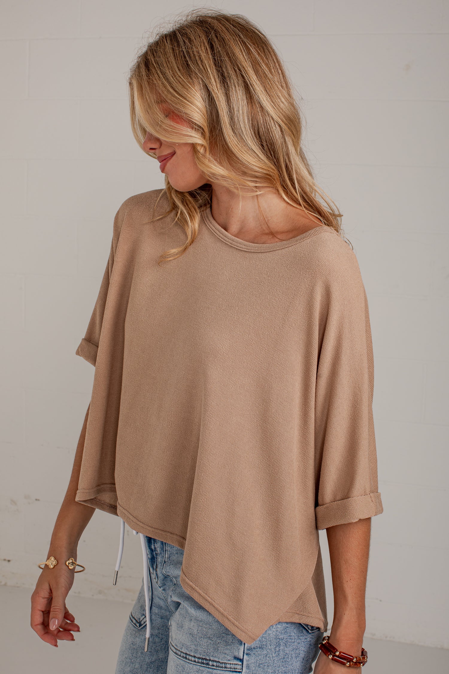 A woman wearing a loose, short-sleeve knit top in a warm taupe-beige color, paired with light-wash blue jeans, standing against a light off-white wall.