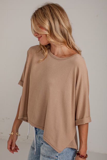 A woman wearing a loose, short-sleeve knit top in a warm taupe-beige color, paired with light-wash blue jeans, standing against a light off-white wall.