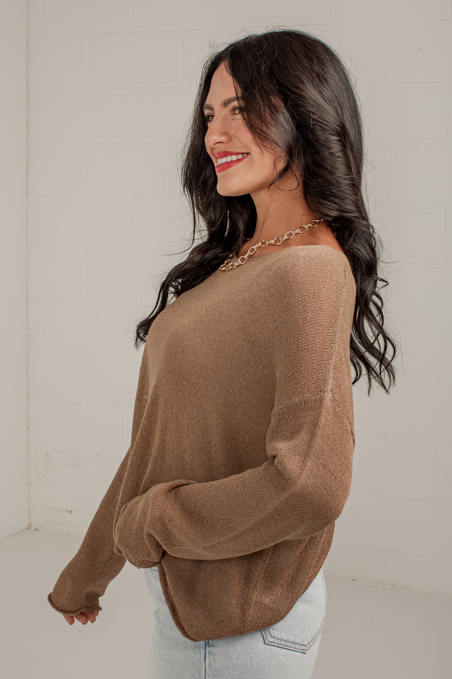 A woman wearing a loose-fitting, long-sleeve knit sweater in a warm taupe-brown color, paired with light-wash jeans and a gold chain necklace, standing against a plain off-white wall.