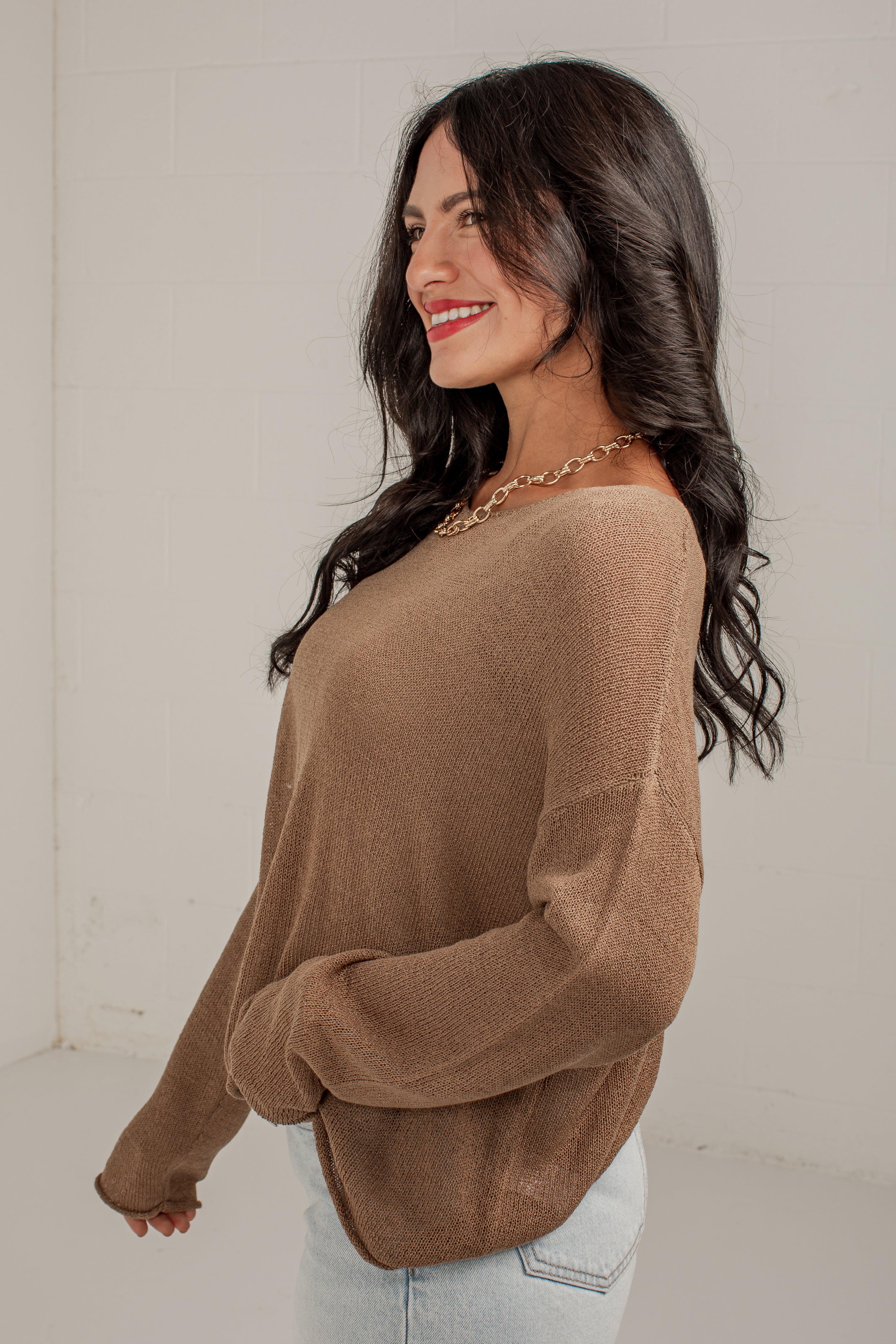 A woman wearing a loose-fitting, long-sleeve knit sweater in a warm taupe-brown color, paired with light-wash jeans and a gold chain necklace, standing against a plain off-white wall.
