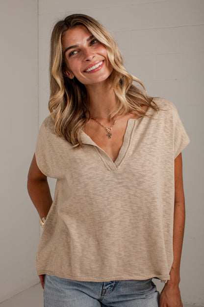 A woman with blonde hair wearing a beige short-sleeve knit top with a split V-neck and blue jeans, standing against a light gray wall.