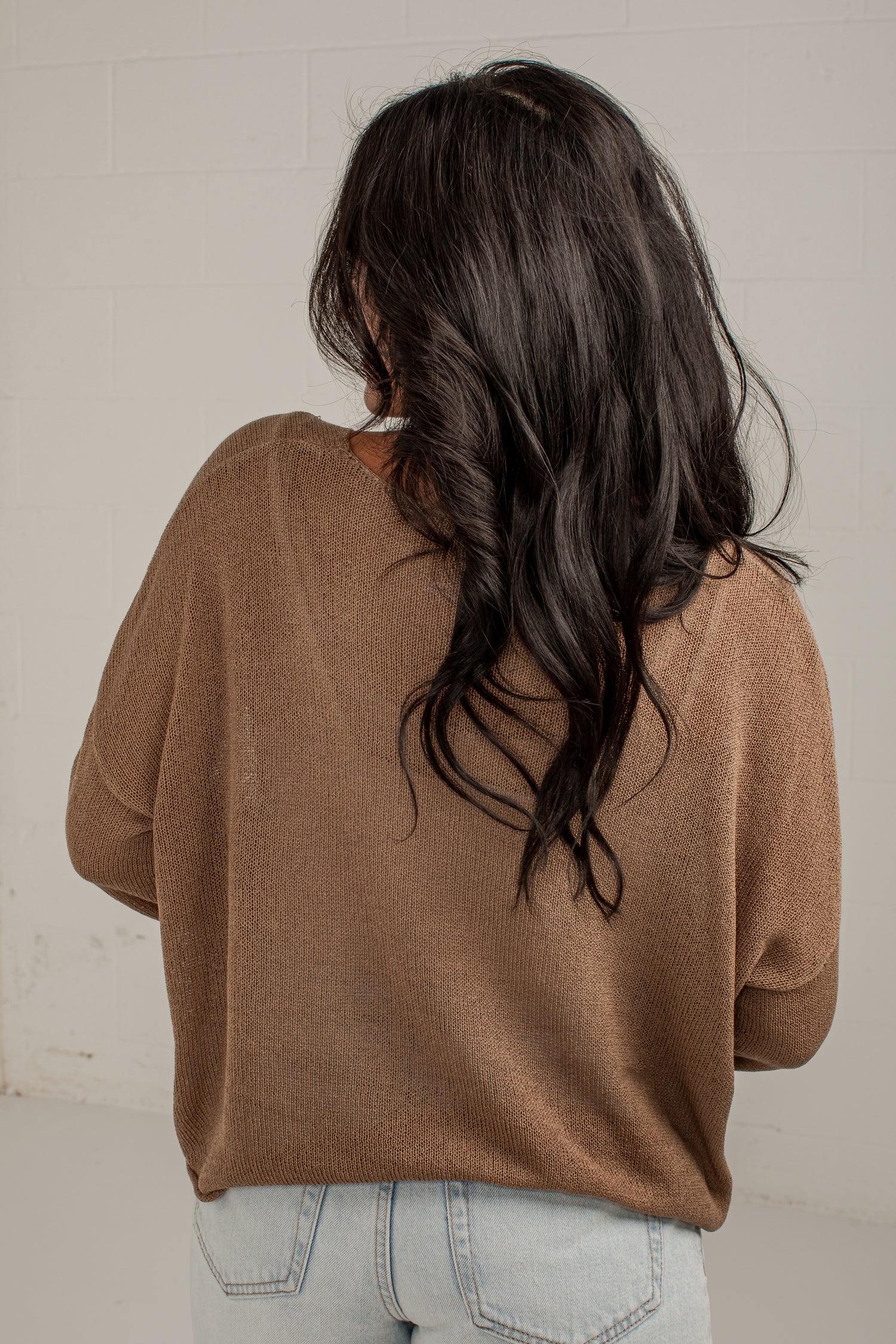 A woman viewed from behind wearing a brown knit sweater and light blue jeans, standing against a white brick wall.