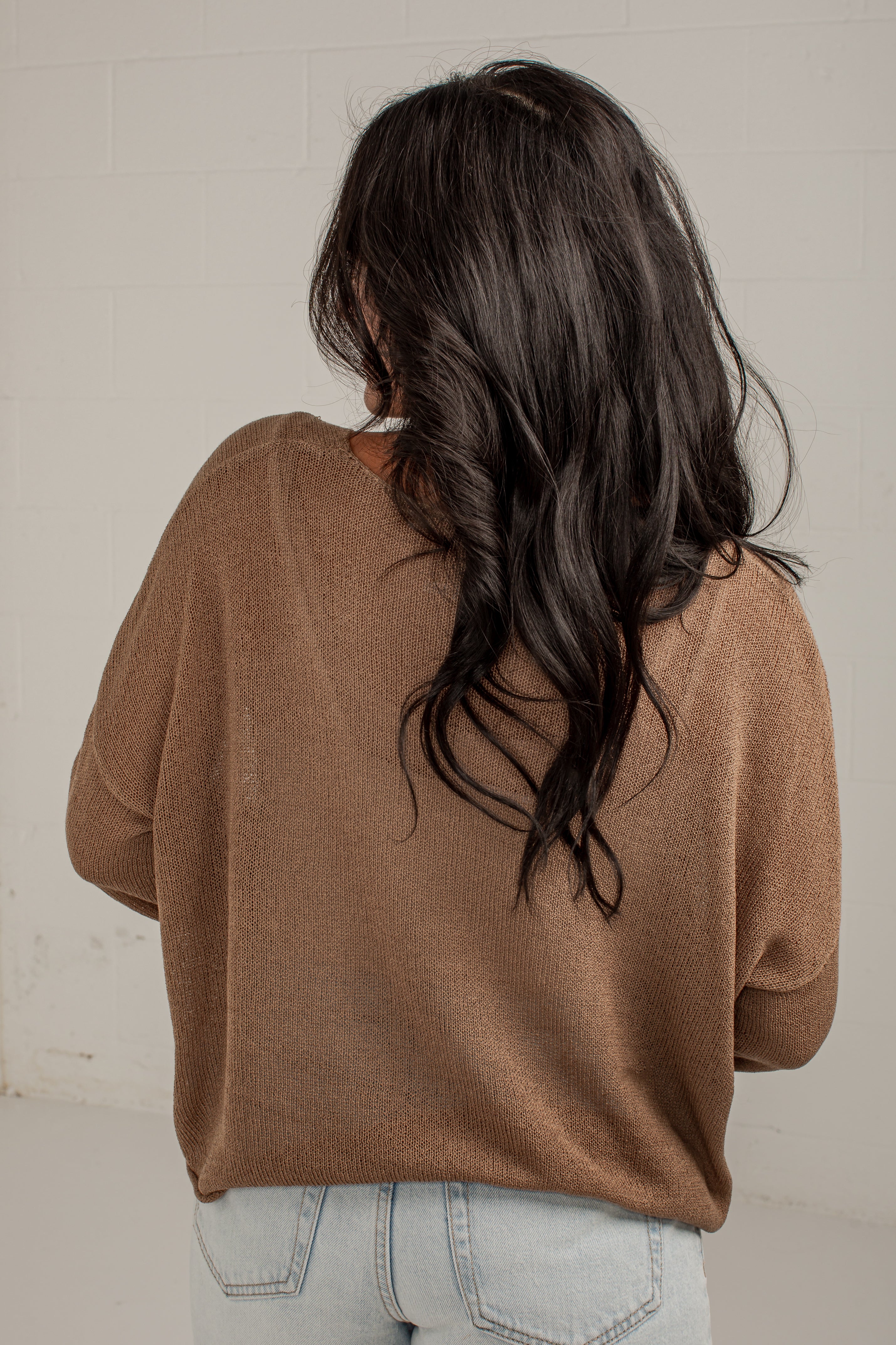 A woman viewed from behind wearing a brown knit sweater and light blue jeans, standing against a white brick wall.