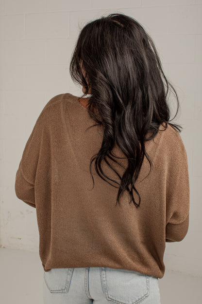 A woman viewed from behind wearing a brown knit sweater and light blue jeans, standing against a white brick wall.