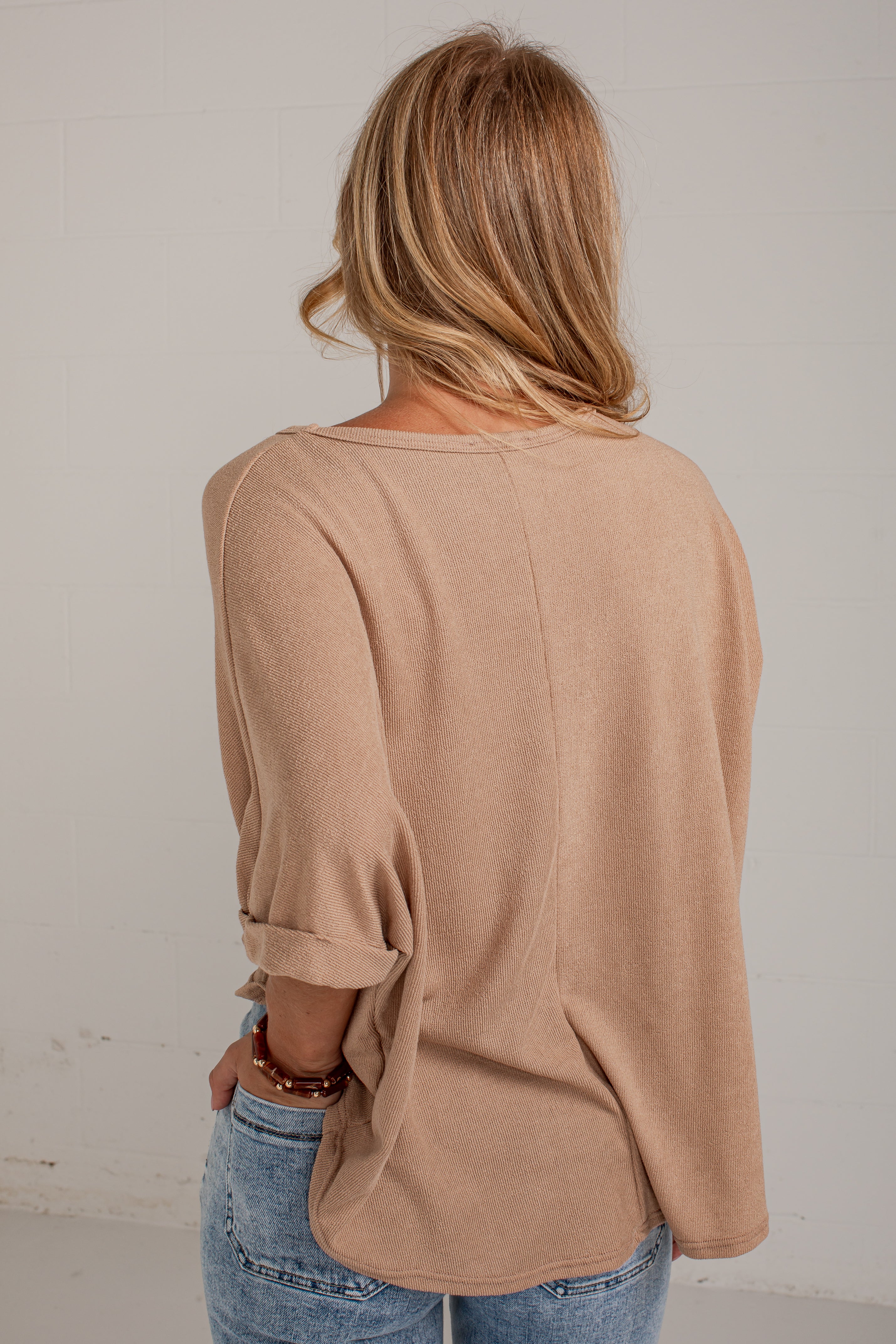A woman viewed from behind wearing a loose, light tan long-sleeve top and blue jeans against a plain off-white wall.