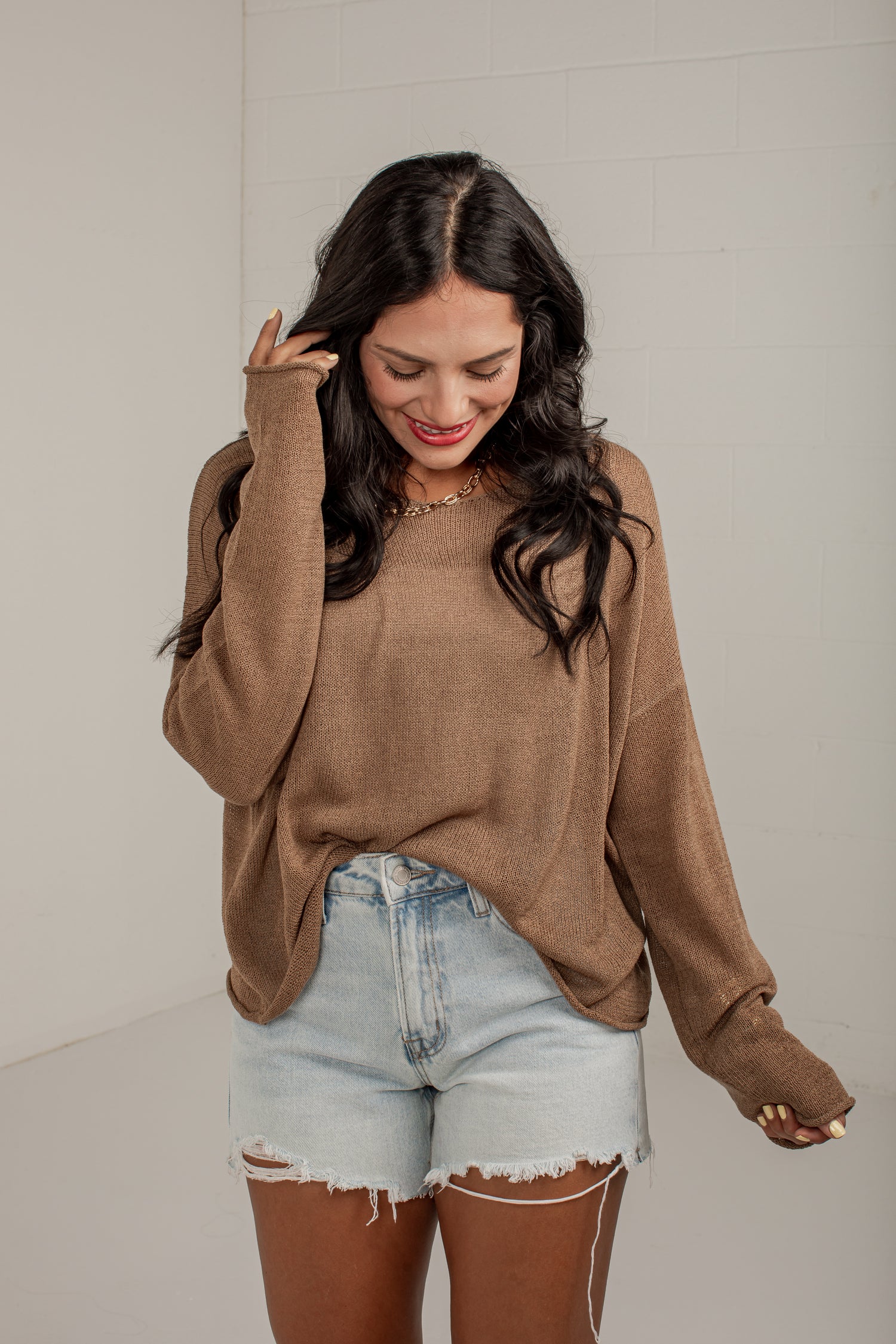 A woman wearing a brown knit long-sleeve sweater and light-wash denim shorts, standing against a light gray wall.