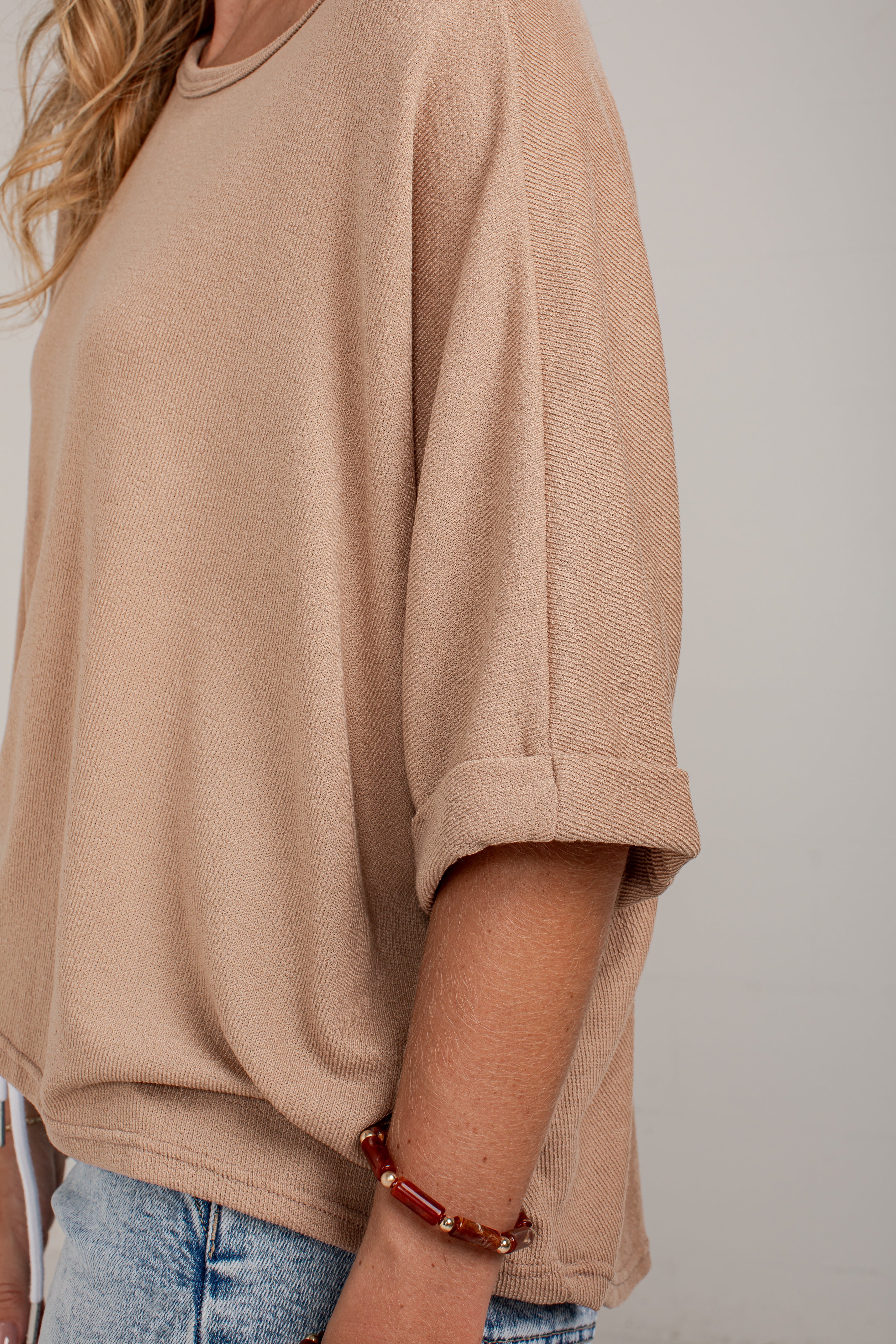 A close-up of a person wearing a loose beige knit top with rolled three-quarter sleeves and light blue jeans against a plain light gray background.