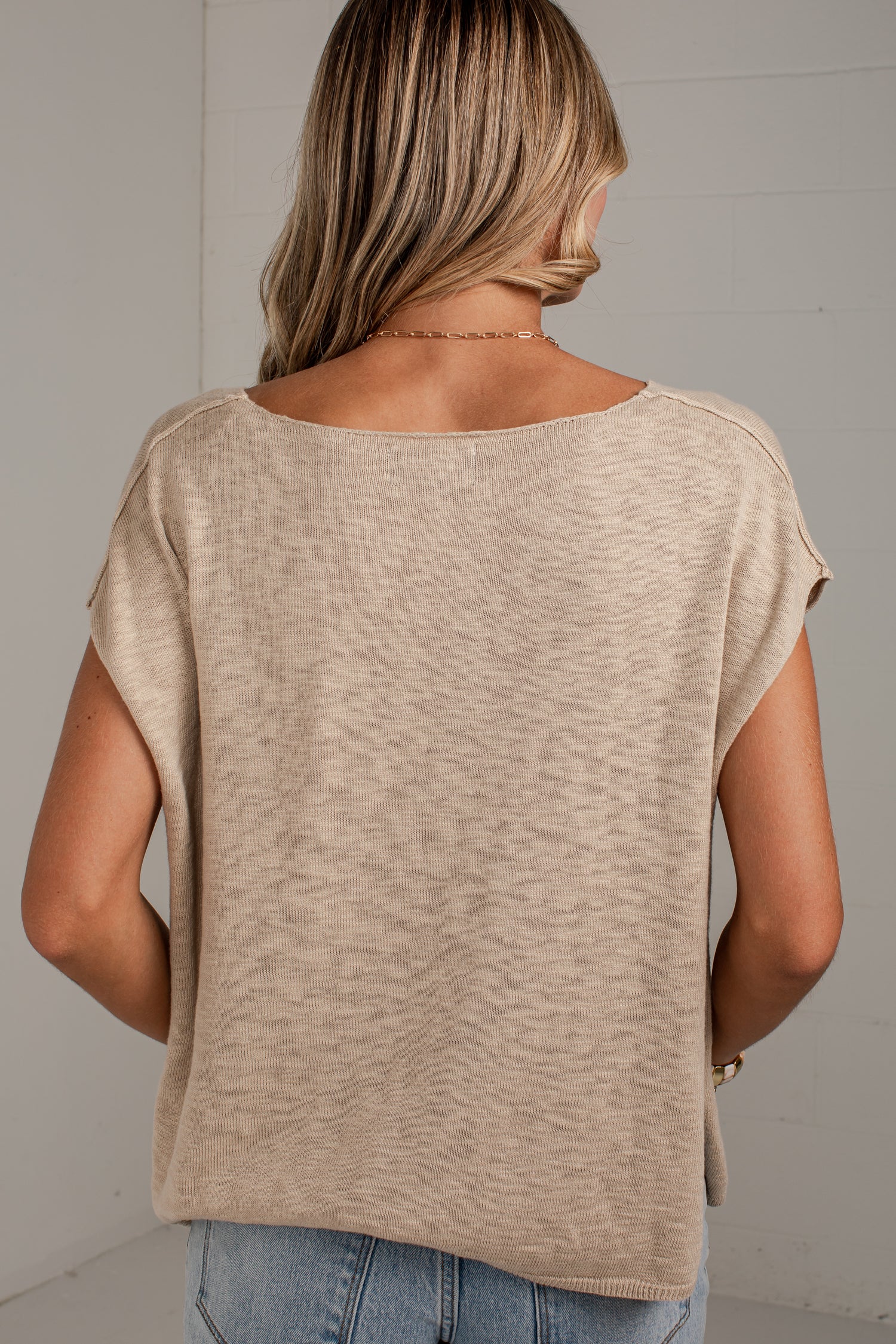 A woman viewed from behind wearing a textured, heathered beige short-sleeve top and light blue jeans against a light gray wall.
