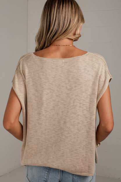 A woman viewed from behind wearing a textured, heathered beige short-sleeve top and light blue jeans against a light gray wall.