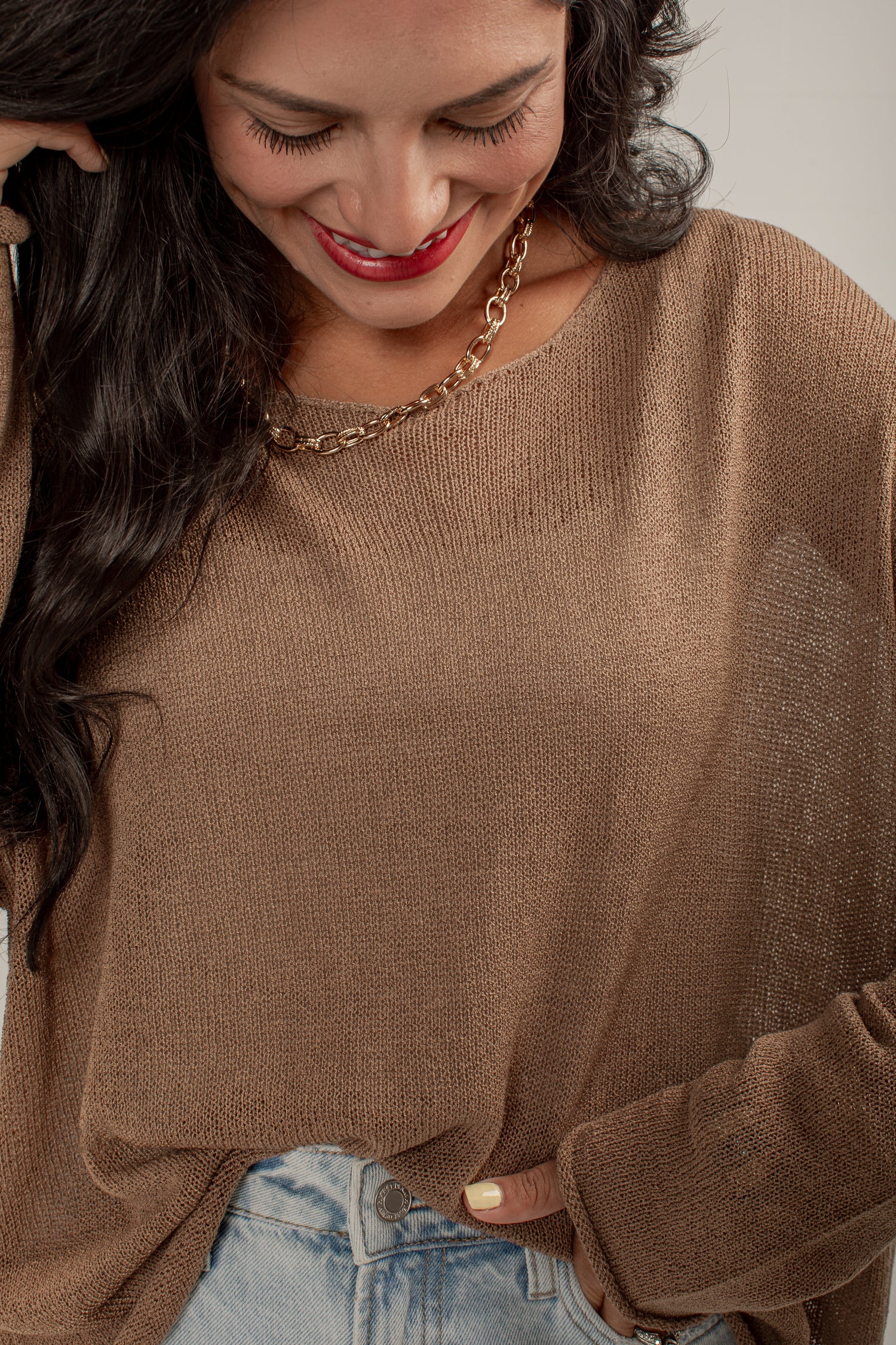 A woman wearing a brown knit long-sleeve top and light blue jeans, shown in a close-up lifestyle shot.