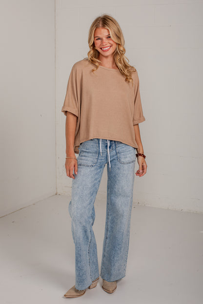A woman modeling a beige short-sleeve cropped top and light-wash blue wide-leg jeans, standing against a plain white studio wall.