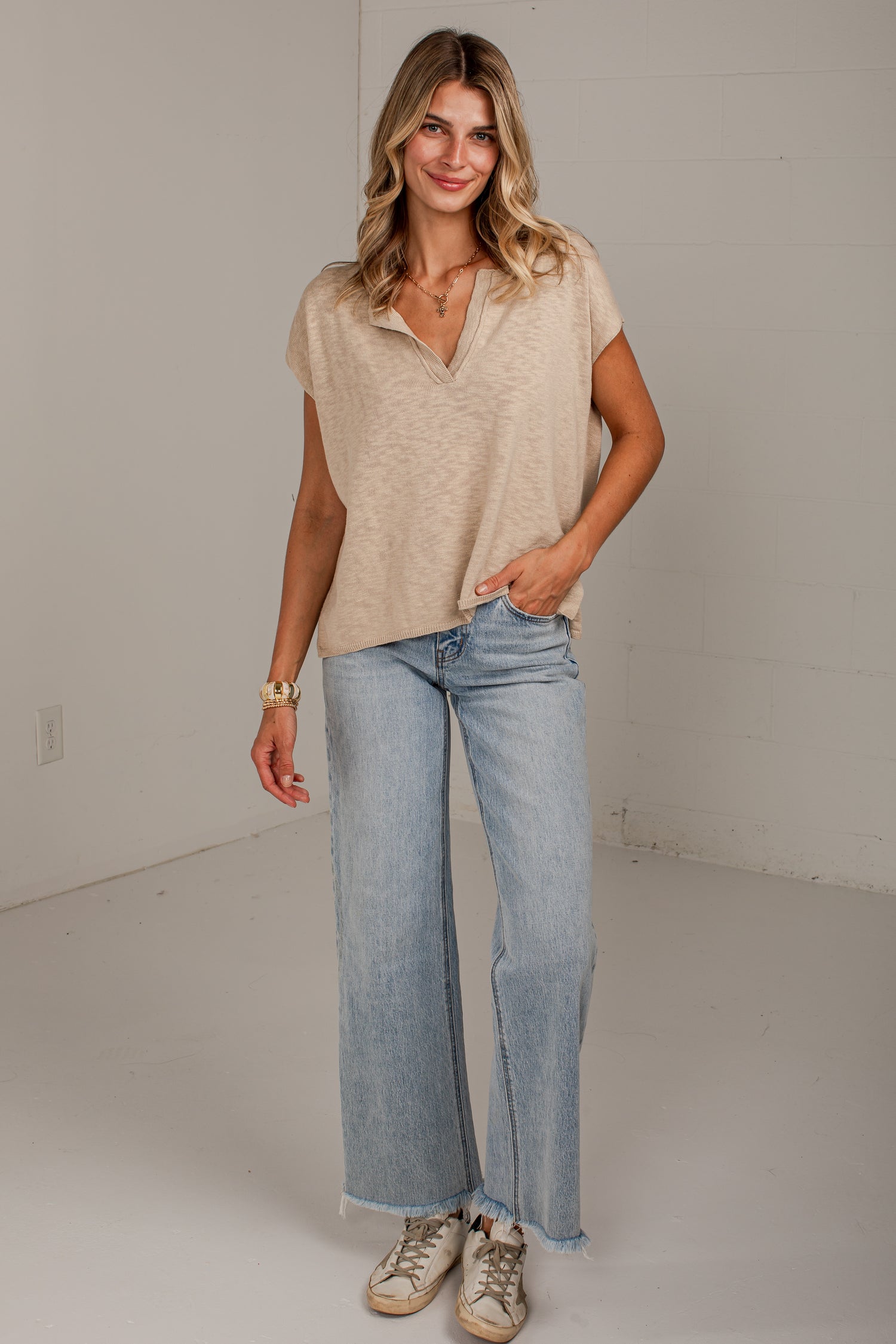 A woman modeling a beige short-sleeve henley-style top and light-wash wide-leg jeans with beige sneakers, standing in a bright studio with a white wall.