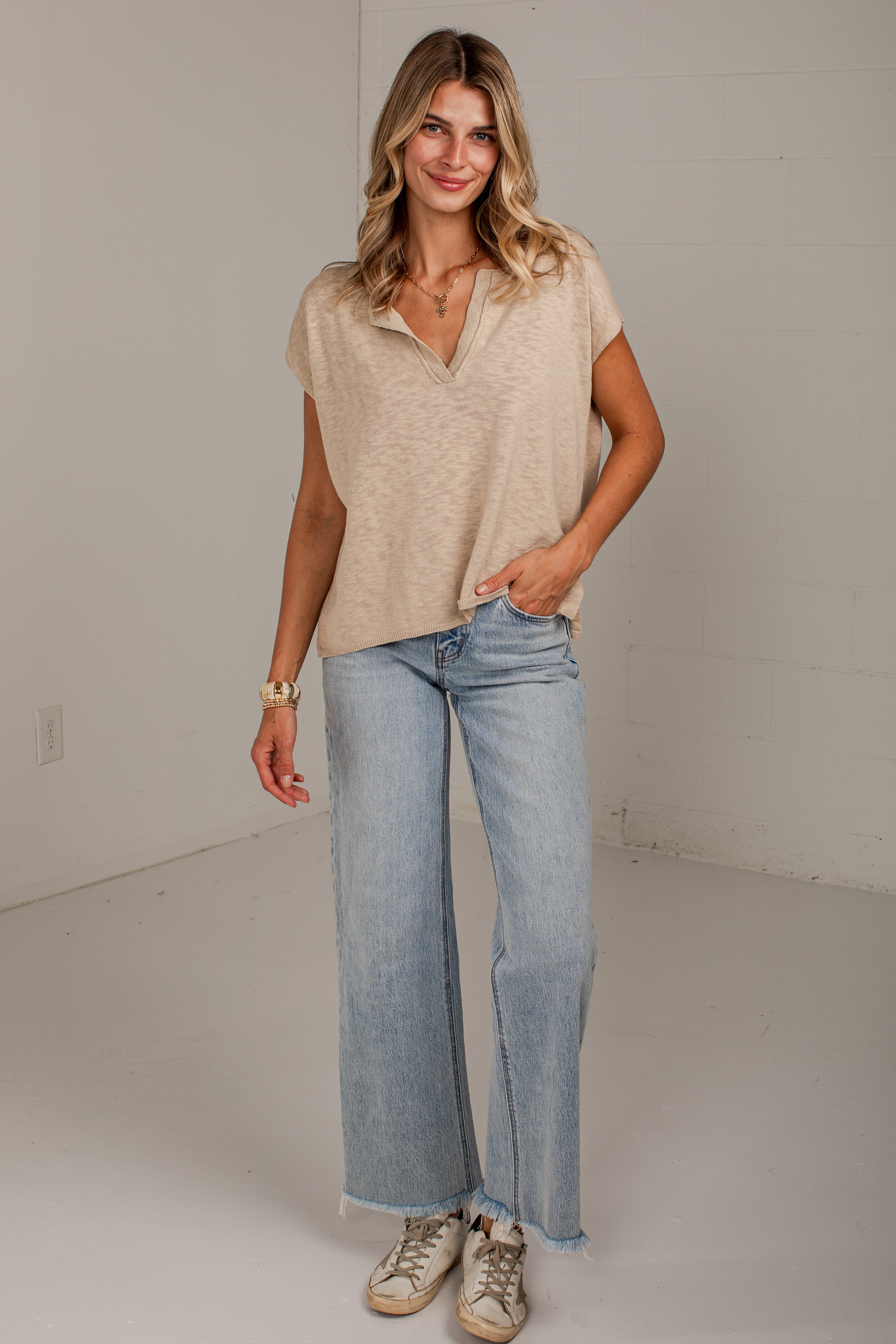 A woman modeling a beige short-sleeve henley-style top and light-wash wide-leg jeans with beige sneakers, standing in a bright studio with a white wall.
