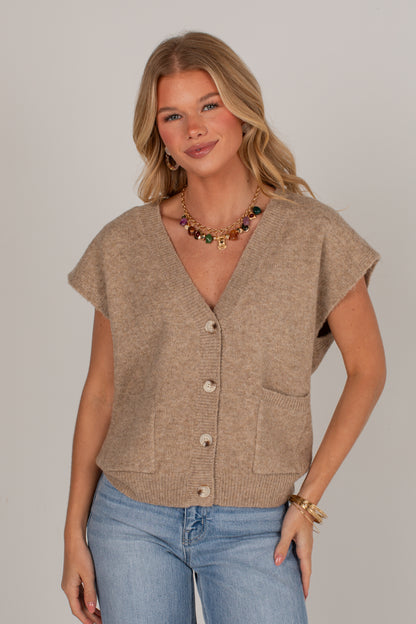 A woman modeling a beige short-sleeve knit cardigan vest with front buttons and patch pockets, paired with light blue jeans, against a solid light gray background.