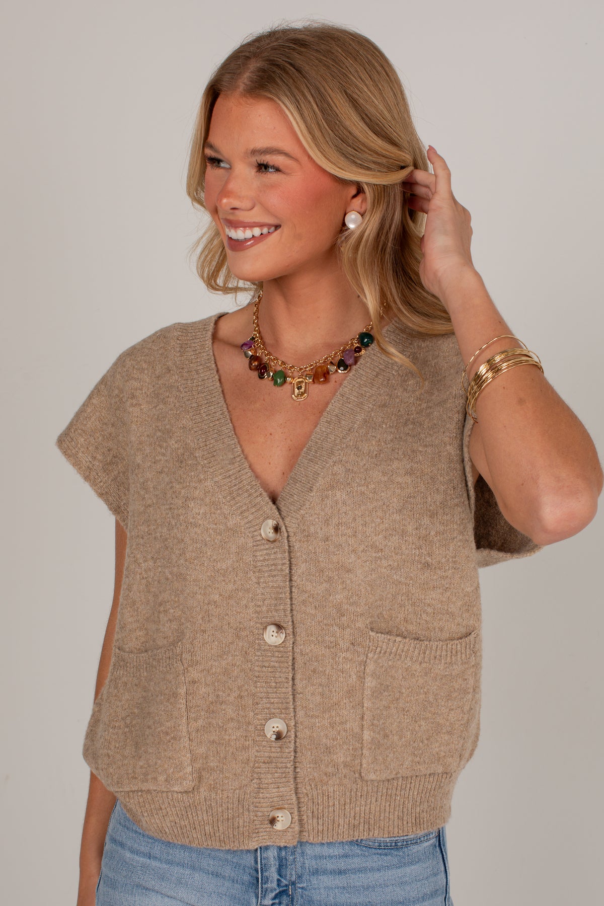 A smiling blonde woman wearing a beige short-sleeve knit cardigan with front buttons and patch pockets, paired with blue jeans, against a plain light gray studio background.