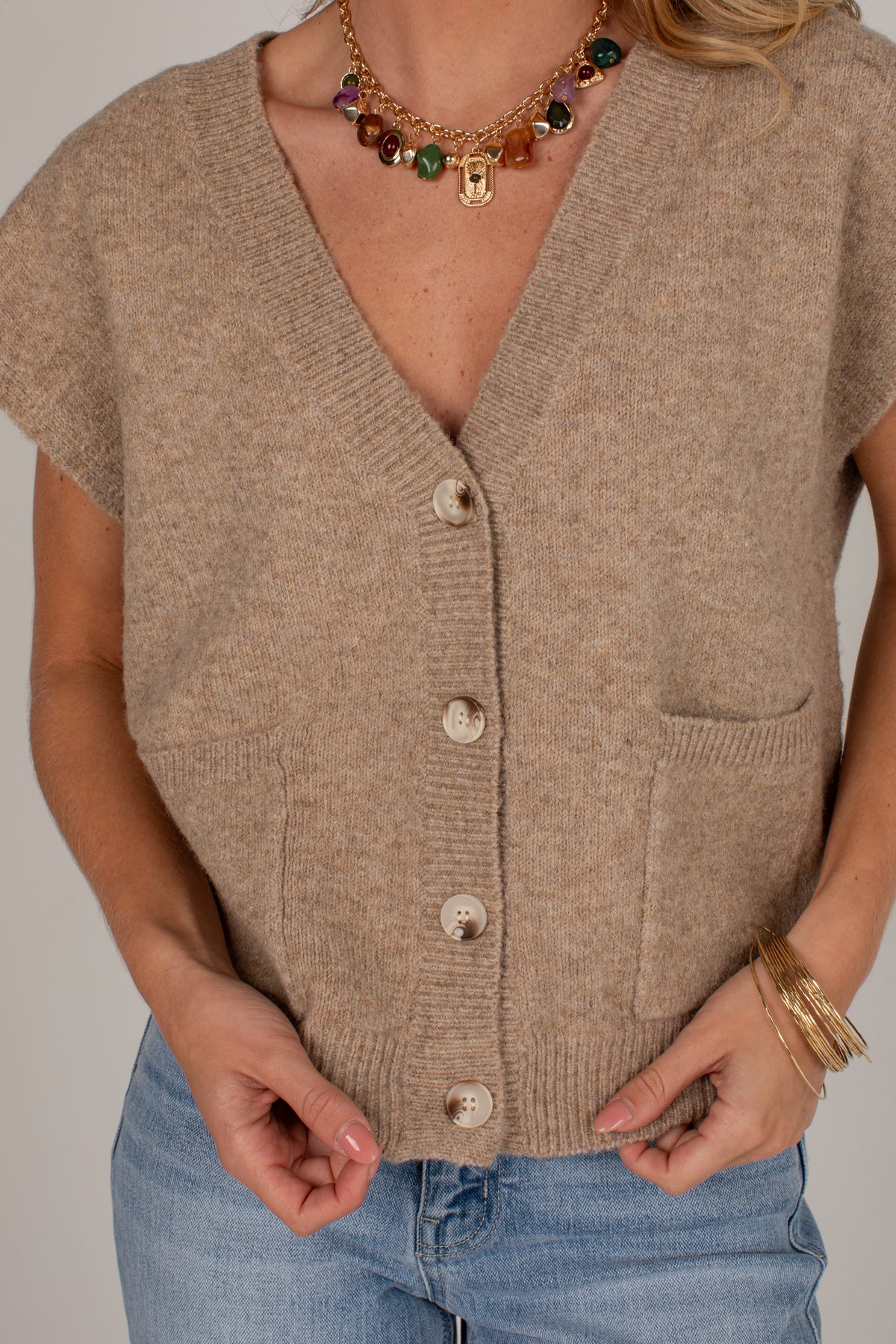 A close-up of a person wearing a beige knit cardigan with short sleeves, a V-neck, front buttons, and a chest pocket, paired with blue jeans. The photo is cropped from the neck to the hips against a plain light background.