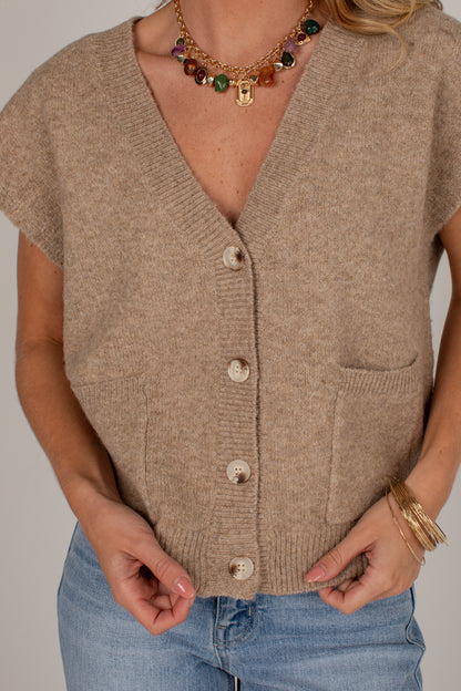 A close-up of a person wearing a beige knit cardigan with short sleeves, a V-neck, front buttons, and a chest pocket, paired with blue jeans. The photo is cropped from the neck to the hips against a plain light background.